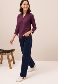 Bordeaux rotes Langarmshirt mit Knopfleiste, kombiniert mit dunkelblauen weiten Jeans und weißen Sneakers mit roten Akzenten. Holzschemel in der Nähe.