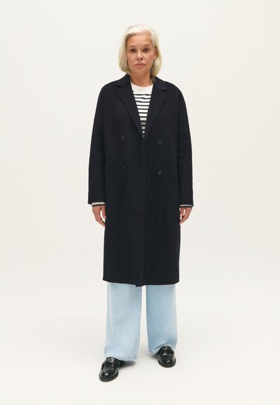Cappotto doppiopetto blu navy con ampi rever e bottoni decorativi, indossato sopra una camicia a righe a maniche lunghe e jeans a gamba larga blu chiaro. Scarpe nere.