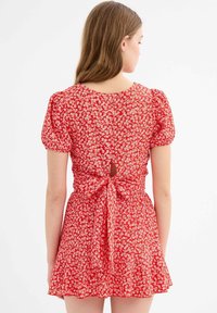 Vestido mini rojo de flores con mangas cortas abullonadas, cintura ceñida y detalle de lazada en la parte posterior. Tejido ligero y texturizado con un patrón divertido.
