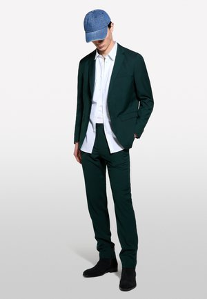 Uomo che indossa un completo verde scuro con camicia bianca, stivali neri e berretto di jeans blu, in piedi con una mano in tasca e testa leggermente inclinata.