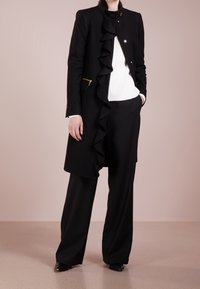 Cappotto in misto lana nera con un ruffle asimmetrico, hardware dorato e colletto alto, indossato sopra un maglione bianco e pantaloni neri.