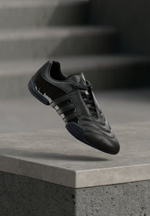 Μαύρο αθλητικό παπούτσι με λείο δέρμα, τρεις αντανακλαστικές ρίγες, υφή στη σόλα και κομψό, στρογγυλεμένο περίγραμμα.