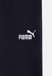 Tumšu zilu audumu ar gludu tekstūru, ar baltu izšūtu Puma logo, kas izvietots pa kreisi. Īso piedurkņu dizains.