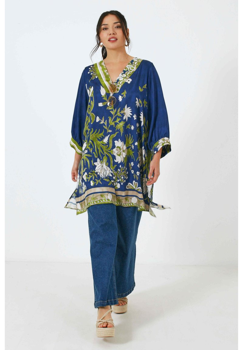Blauwe bloemen tuniek met groene accenten, v-hals en 3/4 mouwen, gecombineerd met lichte denim broeken en plateau sandalen.