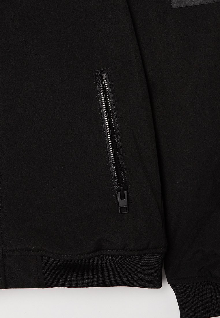 Chaqueta bomber negra con un exterior suave, puños acanalados y un bolsillo con cremallera en la manga izquierda para un almacenamiento seguro.