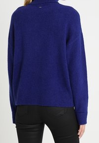 Maglione blu con un alto collo a costine e maniche lunghe e larghe. Tessuto a maglia texturizzato e vestibilità corta con orlo a costine. Pantaloni neri.
