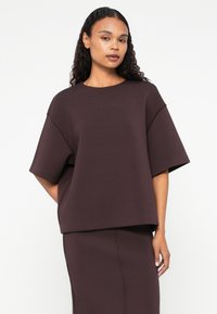 Bordový oversized top vyrobený z hladkej textílie; má okrúhly výstrih, krátke rukávy a voľný strih, v súlade so zodpovedajúcou sukňou.