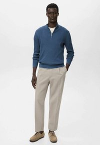 Modra rebrasta jopica s četrtinskim zadrgo, kremna majica, bež hlače in svetlo rjavi čevlji na vezalke. Preprost in sodoben dizajn.