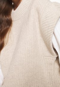 Beige gerippter Strickpullover mit hohem Kragen und kurzen Ärmeln; verfügt über einen strukturierten Stoff und eine lockere Passform.