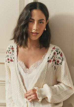 Woman with shoulder-length dark hair wears cream cable-knit cardigan with pink floral embroidery over white lace-trimmed top, standing indoors.