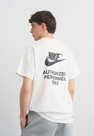 Νέος άνδρας που κοιτάζει αριστερά, φοράει λευκό T-shirt Nike με το κείμενο «Authorized Personnel Only» και γκρι φόρμες.