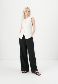 White sleeveless vest with a round neckline and front buttons, paired with loose black trousers and black flip-flops.