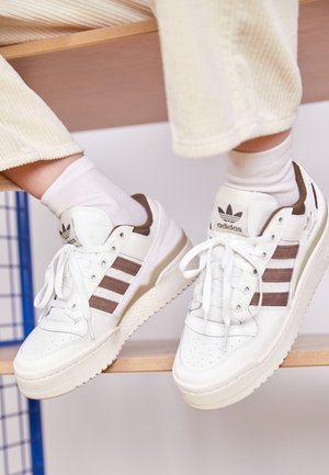 Białe sneakersy Adidas z brązowymi paskami, noszone z białymi skarpetkami i kremowymi spodniami sztruksowymi, stopy spoczywają na drewnianej powierzchni.