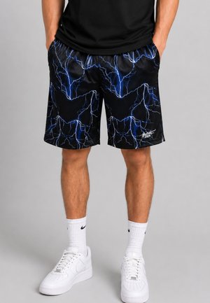 Uomo che indossa pantaloncini neri con motivi a lampo blu, calzini bianchi Nike e scarpe da ginnastica bianche, in piedi con le mani nelle tasche.