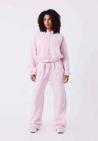 Hoodie zippé rose clair avec une taille ajustée, associé à un pantalon de survêtement assorti à coupe décontractée. Texture lisse et accents de couture décorative.