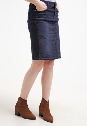 Kvinde iført en mørk denim nederdel til knæene med hænderne i forlommerne og brune ruskindsstøvler med blokhæle.