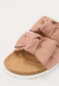 Mīksta rozā zamšādas slaida sandele ar diviem mezglotiem ziediem, korķa zole un balta gumijas zole. Mīksts, teksturēts augšējais slānis ar gludu pamatni.