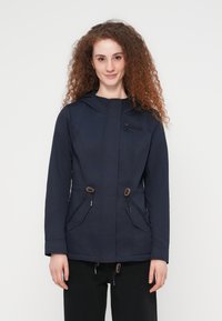 Femme aux cheveux bouclés portant une veste à capuche bleu marine avec des poches devant et une fermeture éclair, debout devant un fond blanc uni.