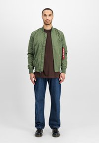 Man staat in een olijfgroene bomberjack, een bruine shirt, blauwe jeans en zwarte schoenen tegen een effen witte achtergrond.