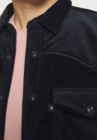 Czarna sztruksowa kurtka na zatrzaski, o fakturowanej tkaninie i kieszeni na piersi. Noszona na jasnoróżowej koszuli, tworząc warstwowy wygląd.