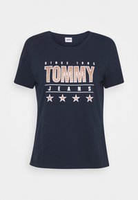 Námořnická modrá bavlněná trička s krátkým rukávem, na kterých je velký potisk "TOMMY JEANS" v metalické bronzové a bílé barvě a pět bílých hvězd.