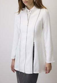 Balta garā piedurkņu jaka ar apkakli, kura priekšpusē ir trīs rāvējslēdzēji un kurai ir mīksta, gluda virsma. Izstrādāta ar piegulošu siluetu un tīrām līnijām.