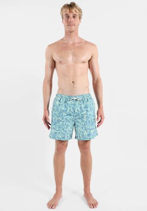 Homme blond debout pieds nus portant un short de bain bleu à motifs avec une ceinture élastique et un cordon, sur un fond uni.
