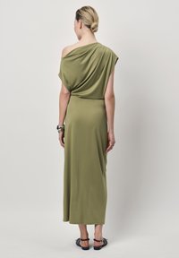 Olijfgroene maxi-jurk met schoudervrije halslijn, gemaakt van zachte stof, met een gedrapeerd ontwerp en een enkellange silhouet, met platte sandalen als schoeisel.