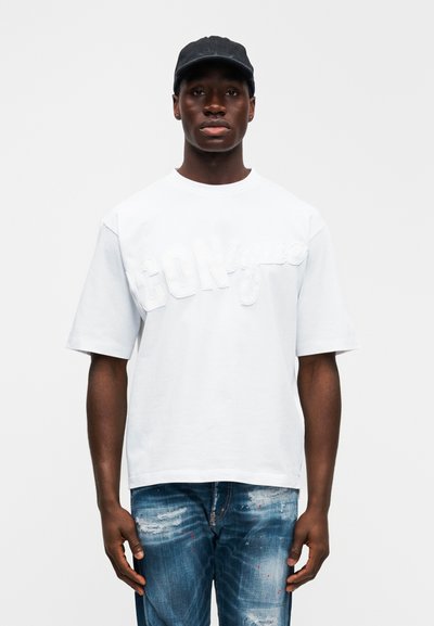 Άσπρο βαμβακερό κοντομάνικο T-shirt με ανάγλυφα γράμματα "ICON", στρογγυλή λαιμόκοψη και κοντά μανίκια. Φορεμένο με μπλε τζιν παντελόνι με φθορές. Απλό φόντο.