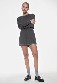 Donkere grijze blouse met lange mouwen en hoge halslijn, gecombineerd met bijpassende shorts met satijnen afwerking. Model draagt grijze sokken en zwarte loafers.