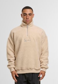 Pulover bej din fleece cu guler cu jumătate de fermoar, manșete ribbate și talie elastică. Prezintă o suprafață texturată și un logo frontal din fir argintiu.