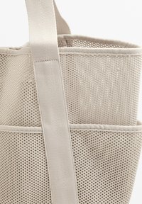 Beige hálós bevásárlótáska textil pántokkal és nyitott tetejével, téglalap alakú szekciókkal és texturált mintával a légáteresztőképesség érdekében.