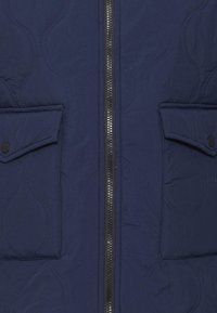Giacca trapuntata blu navy con zip centrale, due tasche frontali con chiusura a bottone e un tessuto liscio e testurizzato.