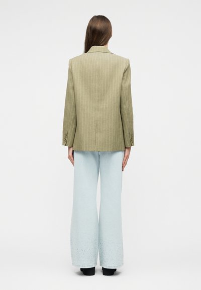 Femme aux longs cheveux bruns portant un blazer à fines rayures vert clair et un jean large bleu clair, debout de dos sur fond blanc.