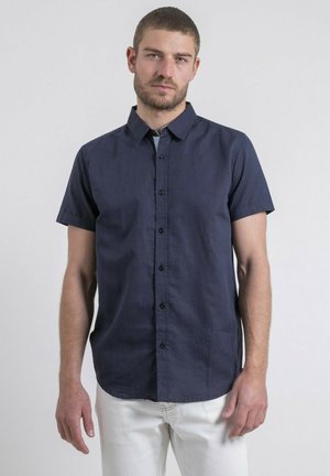 Homme portant une chemise à manches courtes bleu marine à boutons et un pantalon blanc, debout contre un fond clair uni.