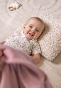 Baby liegt auf einem strukturierten cremefarbenen Kissen mit einem floral gemusterten Overall. Eine pinkfarbene Decke deckt teilweise das Baby zu. Im Hintergrund ein weicher, neutraler Teppich.