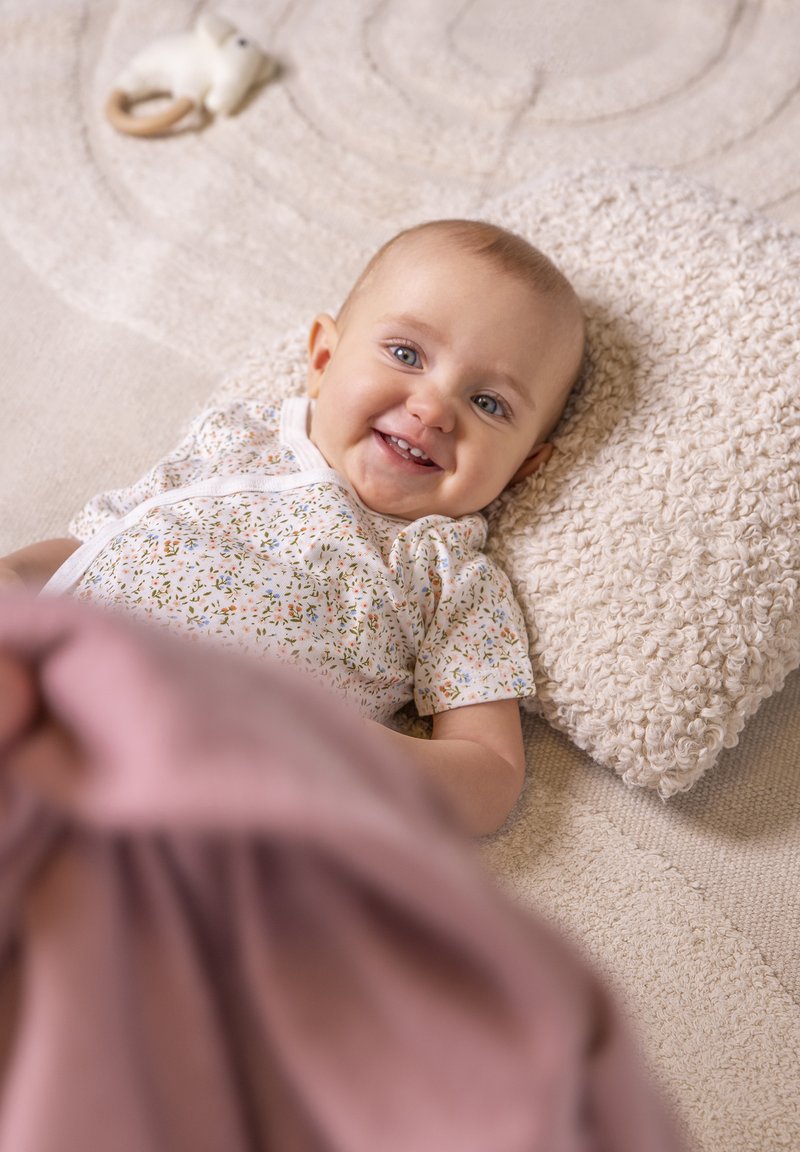 Baby liegt auf einem strukturierten cremefarbenen Kissen mit einem floral gemusterten Overall. Eine pinkfarbene Decke deckt teilweise das Baby zu. Im Hintergrund ein weicher, neutraler Teppich.