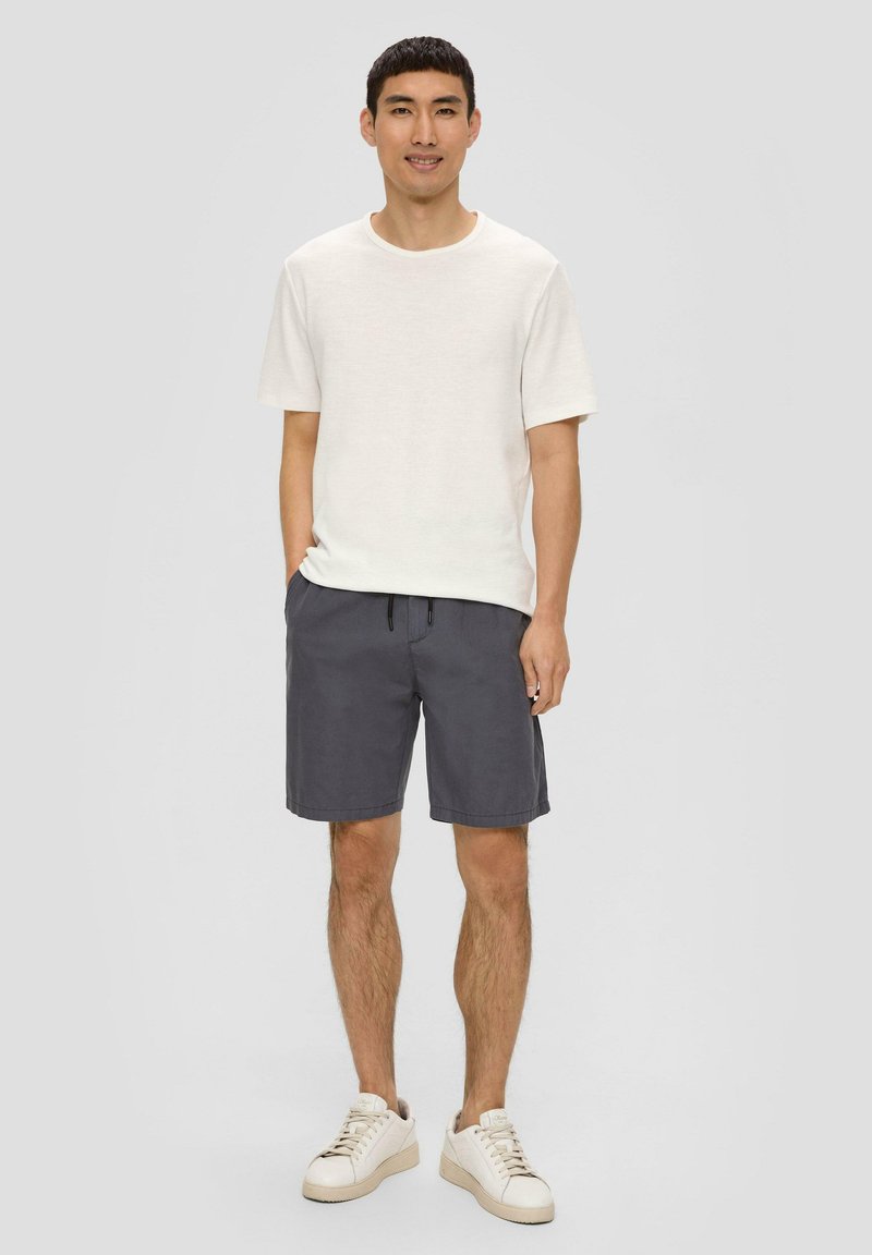 Weißes T-Shirt mit kurzen Ärmeln, graue Shorts mit elastischem Bund und Kordelzug sowie off-white Sneakers, vor einem grauen Hintergrund stehend.