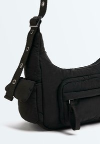Sac bandoulière noir matelassé avec sangle ajustable, poche avant zippée et poche latérale à rabat sur un fond uni.