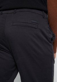 Vue de dos d'une personne portant un pantalon noir Calvin Klein avec une poche passepoilée arrière et un pull noir.