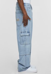 Lys blå cargo jeans med sidelommer og sømdetaljer, med et logo på baklommen. Båret med hvite sneakers.