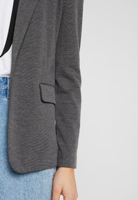 Šedý blazer s texturovanou tkaninou, jednou kapsou na levé straně, spárovaný s bílou košilí a světle modrou džínovou sukní.