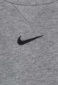 Sudadera gris con cuello en V y ribete acanalado. Presenta un logotipo de Nike bordado en negro en la parte delantera. Tejido suave y texturizado.