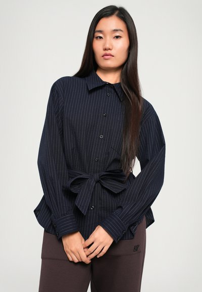 Navy blue striped shirt with long sleeves, button closure, and a tie at the waist. Worn with brown pants. Smooth fabric texture.