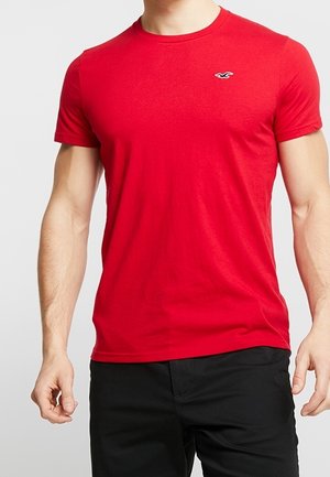 Man draagt een effen rood T-shirt met korte mouwen, een klein wit logo op de linkerborst en zwarte broek.