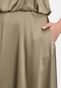Oliwkowa sukienka z teksturowanej tkaniny, z marszczeniem w talii i boczną kieszenią, prezentująca delikatne drapowanie i gładkie wykończenie.