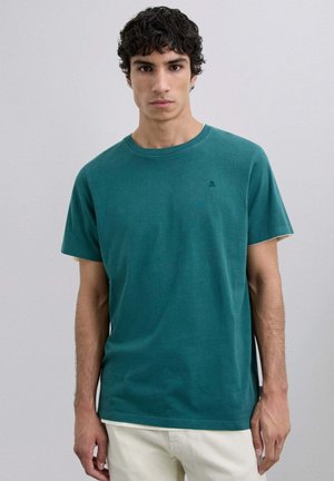 Camiseta de algodón color verde azulado con cuello redondo y mangas cortas, que presenta un pequeño logo en el pecho y un corte relajado.