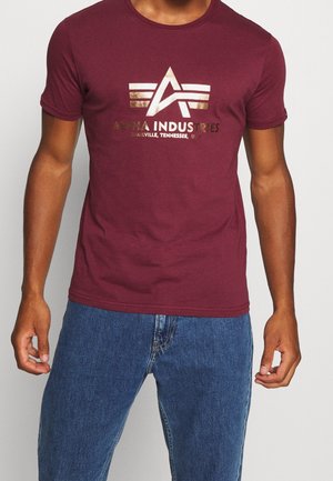 Bordeaux kortærmet t-shirt med guld Alpha Industries-logo og tekst, båret med blå denimjeans på en stående person.