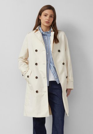 Woman wearing a beige double-breasted trench coat over a blue and white striped shirt and dark blue jeans, standing against a gray background.