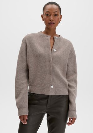 Donna che indossa un cardigan in maglia color taupe con bottoni e pantaloni in pelle nera, in piedi davanti a uno sfondo grigio chiaro uniforme.